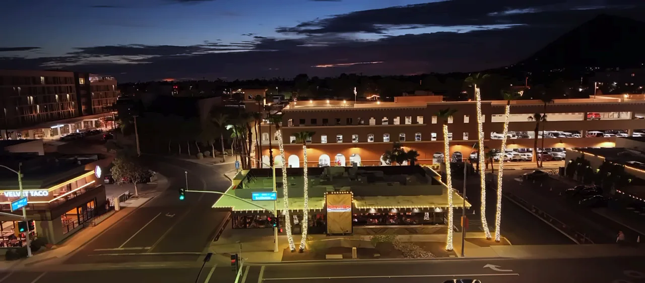 Bourbon & Bones at dusk with light-wrapped palms, seen from a drone above the intersection