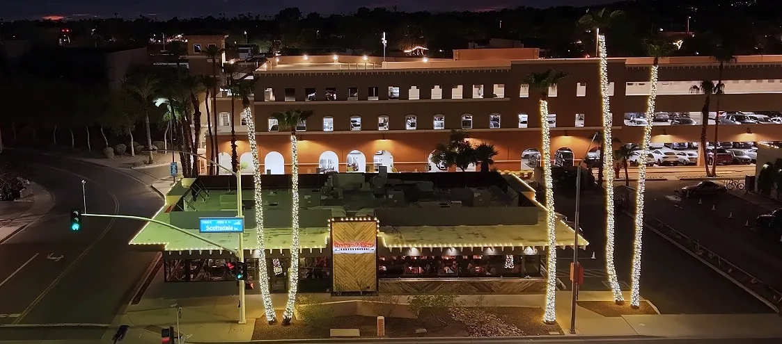 Bourbon & Bones in Scottsdale at night with light-wrapped palm trees and roofline lights, seen from a drone